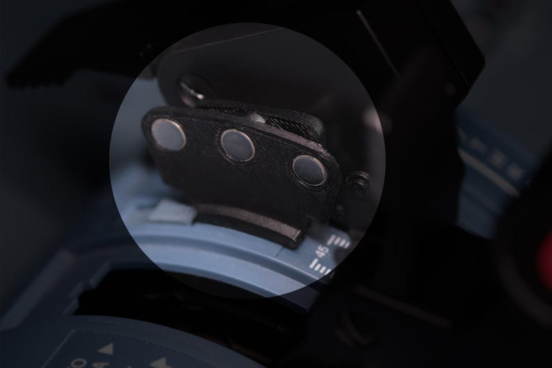 May include: A black, textured, semi-circular component featuring three embedded circular silver magnets. This piece is positioned over a blue, curved mechanical dial with white numerical markings, including the number 45, visible on the surface.