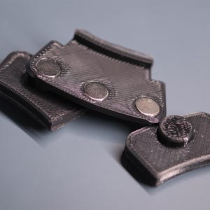 May include: Three black 3D-printed plastic components with a textured, layered surface finish. Each piece features circular metallic magnets embedded into the material, with one piece showing a single magnet and another showing three magnets.
