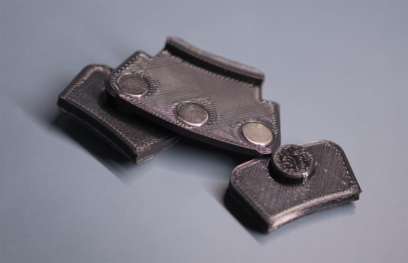 May include: Three black 3D-printed plastic components with a textured, layered surface finish. Each piece features circular metallic magnets embedded into the material, with one piece showing a single magnet and another showing three magnets.