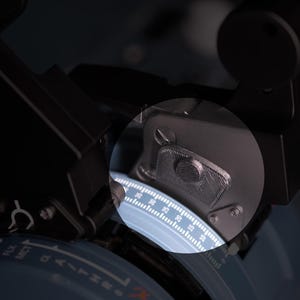 May include: A black flight simulator throttle quadrant featuring a circular dial with white numerical markings and text including TO, GA, FLX, and MCT. A circular magnifying lens highlights a textured metal component and a detailed scale.
