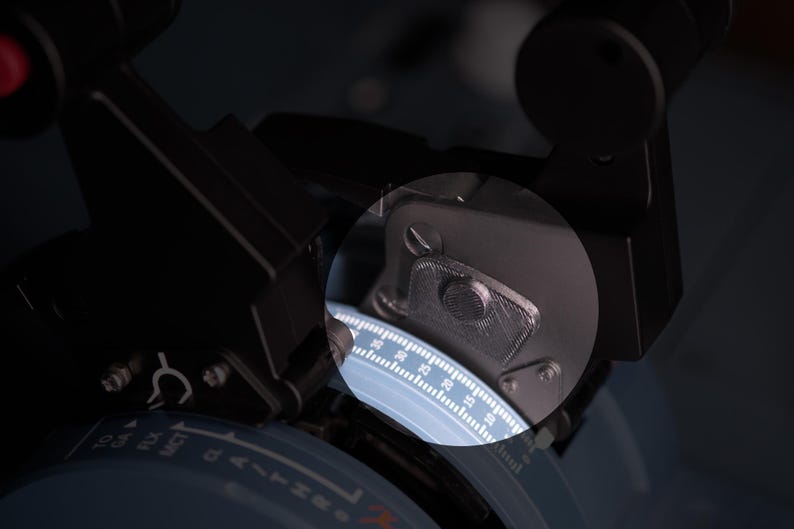 May include: A black flight simulator throttle quadrant featuring a circular dial with white numerical markings and text including TO, GA, FLX, and MCT. A circular magnifying lens highlights a textured metal component and a detailed scale.
