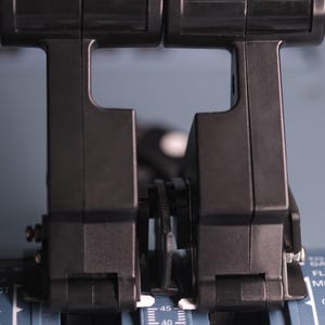 May include: Two black throttle levers positioned side-by-side on a grey control panel. The levers feature a vertical design with integrated triggers. The base panel displays white text markings including GA, FLX, MCT, and CL, along with numerical scales.