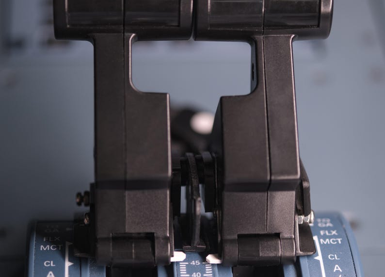 May include: Two black throttle levers positioned side-by-side on a grey control panel. The levers feature a vertical design with integrated triggers. The base panel displays white text markings including GA, FLX, MCT, and CL, along with numerical scales.