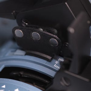 May include: A black mechanical component with three circular silver magnetic points attached to a blue base featuring white numerical markings and a scale. The device includes a black lever arm and a textured surface, showing precise engineering details.