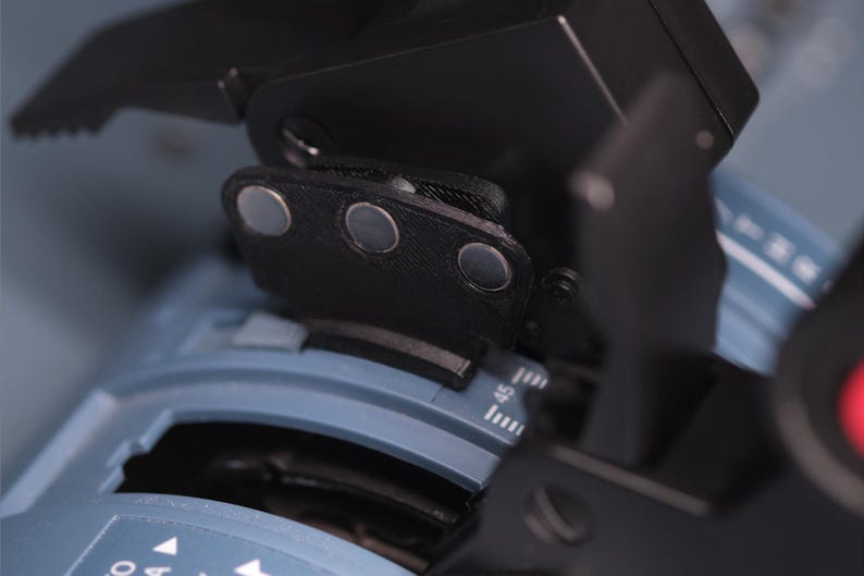 May include: A black mechanical component with three circular silver magnetic points attached to a blue base featuring white numerical markings and a scale. The device includes a black lever arm and a textured surface, showing precise engineering details.