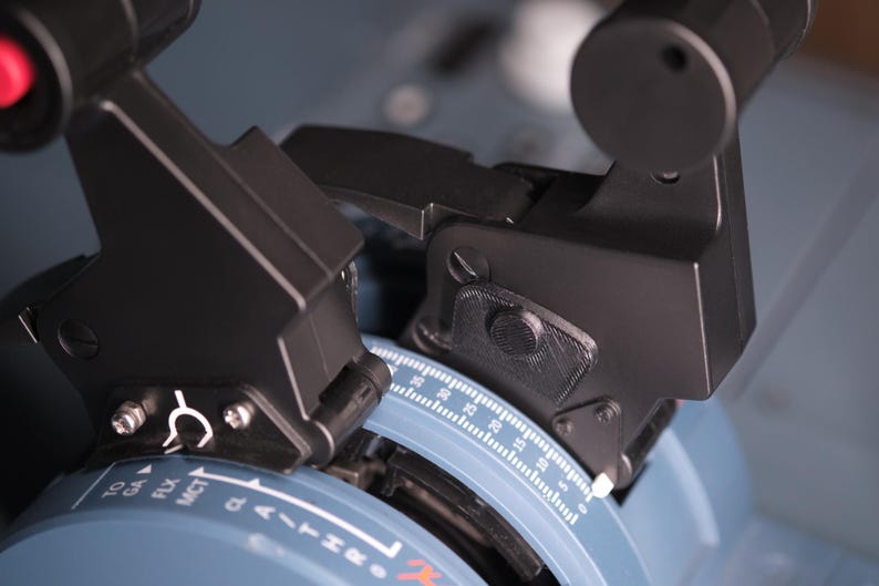 May include: A flight simulator throttle quadrant featuring two black levers with textured grips and a red button. The base is light blue with white printed markings including TO, GA, FLX, MCT, and a numerical scale ranging from 0 to 30.