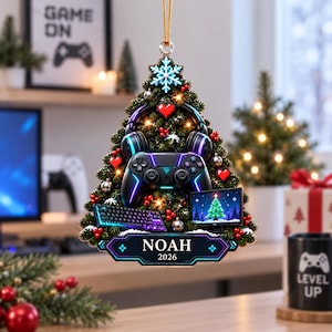 May include: A triangular Christmas tree ornament featuring a gaming controller, headphones, a keyboard, and a monitor. The ornament is decorated with red berries, silver baubles, and a blue snowflake. A plaque at the base displays the text NOAH 2026.