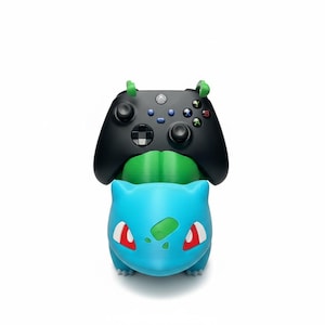 May include: A black wireless video game controller resting on a blue and green creature-shaped plastic stand. The controller features a central Xbox logo, two analog sticks, a directional pad, and four colored action buttons labeled A, B, X, and Y.