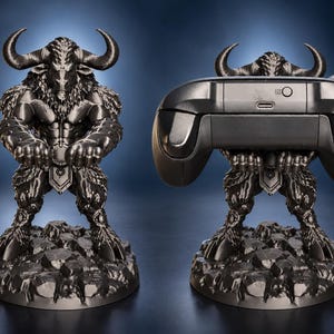 May include: A dark metallic gray figurine of a minotaur standing on a rocky base with its arms extended forward. The figure is designed to hold a black video game controller, which is shown resting in the minotaur's hands in the second view.