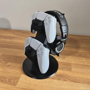 May include: A black plastic gaming accessory stand featuring a circular base, a vertical support column, and integrated mounts holding two white and black wireless game controllers and a pair of black over-ear headphones with white logo accents.