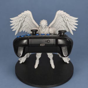 May include: A white figurine of a winged humanoid character standing on a black circular base, holding a black video game controller with blue buttons. The character features large, detailed feathered wings extending outward.
