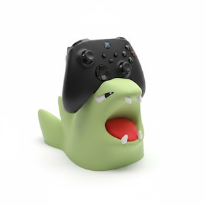 Puede incluir: Un mando de videojuego inalámbrico negro sobre un soporte de color verde claro con forma de criatura. El soporte presenta una boca abierta con una lengua roja, dos dientes blancos y ojos entrecerrados para sujetar el mando.