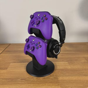 May include: A black vertical stand holding two purple wireless game controllers and a pair of black over-ear headphones. The controllers feature standard button layouts with colored A, B, X, and Y buttons. The headphones have a black headband.