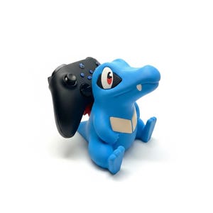 以下が含まれることがあります： 黒いワイヤレスゲームコントローラーを保持する、ワニのような青い生き物のフィギュア型コントローラースタンド。この生き物は、クリーム色の胸の模様、赤い瞳の大きな目、そして口の中に一本の白い歯が見えるデザインが特徴です。