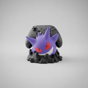 May include: A purple Gengar character figure serving as a controller stand, featuring red eyes and a wide white grin, emerging from a black base. A black wireless gaming controller is positioned behind the figure, resting on the stand.