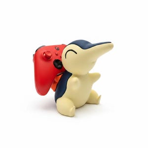 May include: A cream and dark blue creature figurine designed as a controller stand, featuring a smooth matte finish and a rounded, seated posture. A bright red wireless gaming controller is positioned securely against the back of the figure.