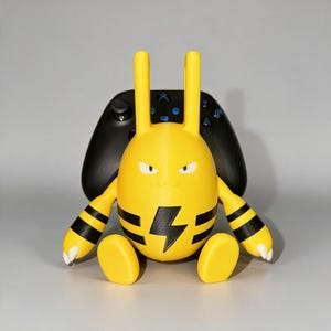 May include: A yellow, egg-shaped character figure with two long ears, white eyes, and a black lightning bolt emblem on its chest. The figure has black bands on its arms and is positioned in front of a black video game controller used as a stand.