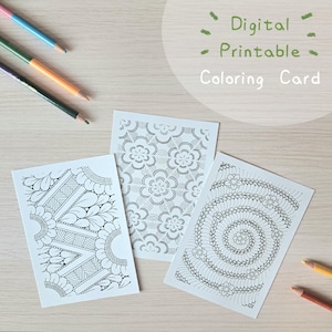May include: Three rectangular white cards featuring intricate black line art patterns, including floral motifs, geometric shapes, and a spiral design. The text Digital Printable Coloring Card is visible in green. Several colored pencils are placed nearby.