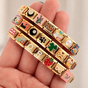 Könnte beinhalten: Drei goldfarbene modulare Gliederarmbänder mit verschiedenen bunten Emaille-Anhängern, darunter eine Acht-Kugel, eine schwarze Katze, eine Erdbeere, ein Kleeblatt, eine Perle, Bücher, ein Herz, die Buchstaben O und R sowie der Text you are worth it.