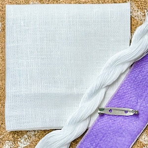 May include: A craft kit with a white linen square, white embroidery floss, a purple ribbon, and a silver pin back. The items are arranged on a corkboard surface, suggesting a DIY project or craft supply set.