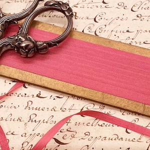 May include: Close-up of antique silver scissors with floral detailing resting on a pink ribbon and a gold-edged card. The background features handwritten script on aged paper, creating a vintage aesthetic.
