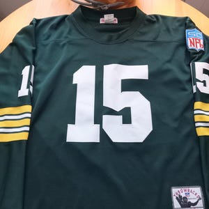 May include: A dark green long-sleeve football jersey featuring the white number 15 on the chest and sleeves, with yellow and white stripes on the cuffs. It includes an NFL 50th anniversary patch, a Mitchell & Ness tag, and a 1969 Bart Starr label.