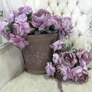 May include: A bouquet of purple silk flowers in a brown pot with a decorative design. The flowers are arranged in a vase on a white tufted chair.