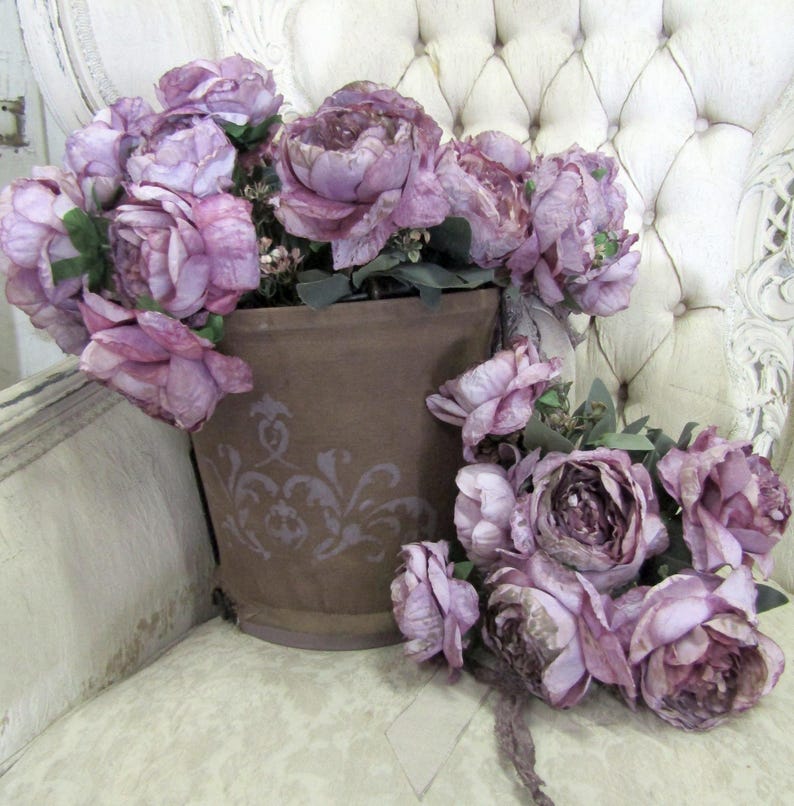 May include: A bouquet of purple silk flowers in a brown pot with a decorative design. The flowers are arranged in a vase on a white tufted chair.
