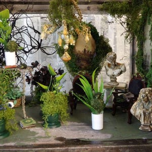 Op de afbeelding: Een miniatuur diorama met diverse planten, mos en decoratieve elementen. Bevat kleine bustes, een miniatuurtafel en een stoel. De sc&egrave;ne is ingesloten in een glazen doos, waardoor een grillige, artistieke weergave ontstaat.