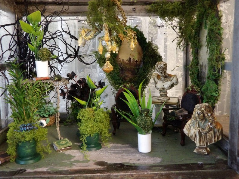 Op de afbeelding: Een miniatuur diorama met diverse planten, mos en decoratieve elementen. Bevat kleine bustes, een miniatuurtafel en een stoel. De sc&egrave;ne is ingesloten in een glazen doos, waardoor een grillige, artistieke weergave ontstaat.