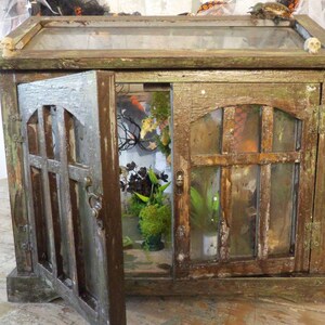 Op de afbeelding: Een verweerde houten kast met een glazen bovenkant en deuren, waarvan er &eacute;&eacute;n open is. De kast heeft een verweerde, verouderde uitstraling met groene en bruine tinten. Binnenin zijn planten en decoratieve elementen zichtbaar. Kleine schedelaccenten zitten bovenop.