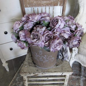 May include: A bouquet of purple silk peonies in a rustic metal bucket with a decorative design. The bucket is sitting on a distressed white wooden stool.