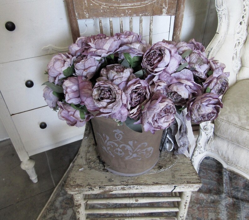 May include: A bouquet of purple silk peonies in a rustic metal bucket with a decorative design. The bucket is sitting on a distressed white wooden stool.