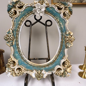 May include: An ornate, oval picture frame with a teal and gold finish. The frame features grape cluster and floral detailing, and is supported by a dark metal easel. A white flower adorns the top.