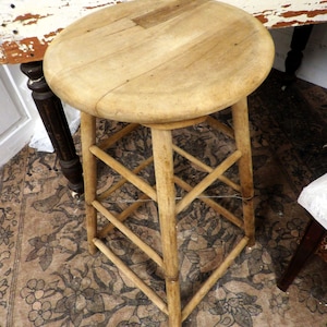 May include: A light-colored wooden stool with a round seat and a crisscross leg design. The stool is made of unfinished wood, showcasing the natural grain. The stool is a classic piece of furniture.