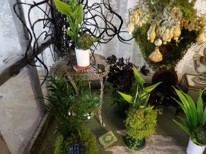 Op de afbeelding: Een interieur met potplanten en decoratieve elementen. Een kleine, sierlijke metalen tafel draagt een witte pot met groen en een miniatuurboek. Andere planten zijn met mos bedekte potten en een groot bloemstuk.