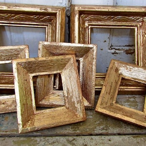 May include: A collection of six antique gold picture frames. The frames have ornate detailing and a distressed finish, with square openings. The frames are arranged on a weathered wooden surface, showcasing their vintage aesthetic.