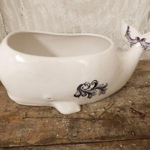 May include: White ceramic whale-shaped planter with a wide opening. The whale has a tail and a fin, both decorated with a dark blue floral design. The planter is on a wooden surface.