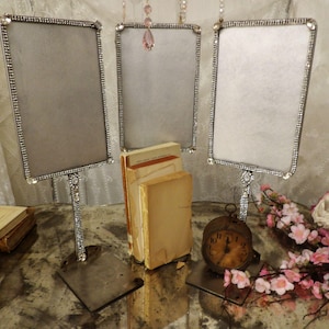 May include: Three silver-framed display stands with rectangular openings, embellished with rhinestones. The stands are on a reflective surface, with old books and a vintage clock. Pink flowers and a white rose are in the background.