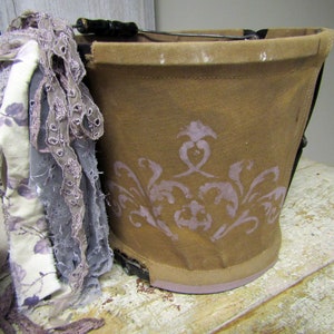 May include: A brown canvas bucket with a purple floral design. The bucket has a black metal handle and is decorated with a white and purple lace trim.
