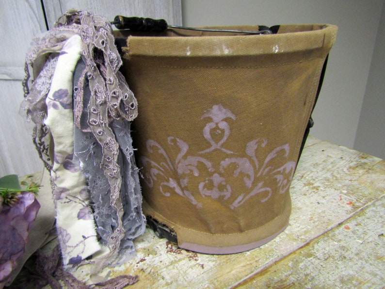 May include: A brown canvas bucket with a purple floral design. The bucket has a black metal handle and is decorated with a white and purple lace trim.