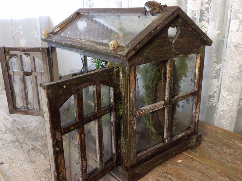 Op de afbeelding: Een rustiek houten terrarium met glazen ramen en een scharnierende deur. Het terrarium heeft een puntdak met een hartvormige uitsparing en is versierd met kleine schedelvormige decoraties. Bovenop zit een kikker.