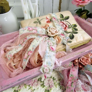 May include: A pink, distressed wooden tray holds a floral-decorated book, ribbons, and fabric. The book has a cream-colored cover with a rose illustration. Ribbons and fabric feature pink and blue floral patterns, with a large bow and rose accents.
