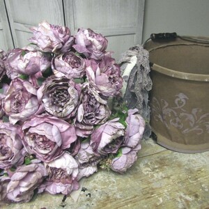 May include: A bouquet of purple and white silk flowers arranged in a brown bucket with a floral design. The bucket has a metal handle.
