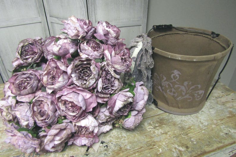 May include: A bouquet of purple and white silk flowers arranged in a brown bucket with a floral design. The bucket has a metal handle.
