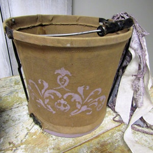 May include: A brown canvas bucket with a metal handle and a floral design. The bucket is sitting on a wooden table with a white lace ribbon.