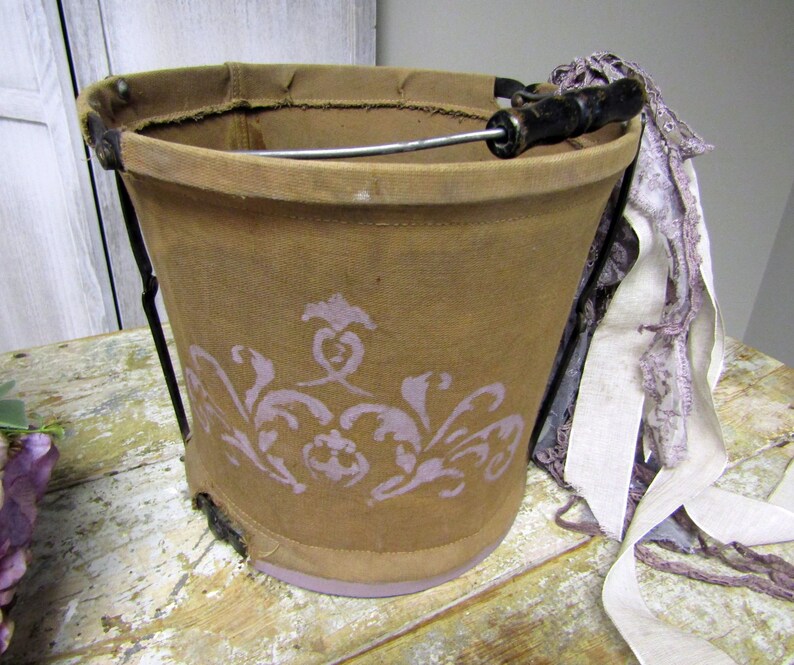 May include: A brown canvas bucket with a metal handle and a floral design. The bucket is sitting on a wooden table with a white lace ribbon.
