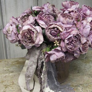 May include: A bouquet of dusty pink peonies arranged in a brown ceramic vase. The peonies are surrounded by a white and grey lace fabric.