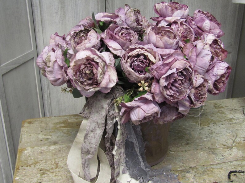 May include: A bouquet of dusty pink peonies arranged in a brown ceramic vase. The peonies are surrounded by a white and grey lace fabric.