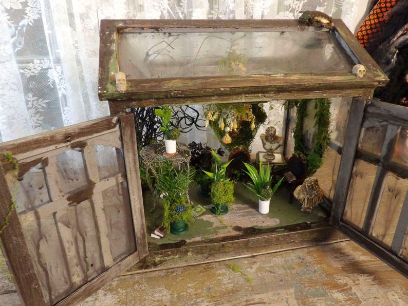 Op de afbeelding: Een miniatuur terrarium in een hergebruikt houten raamkozijn. De binnenkant bevat kleine planten, mos en decoratieve elementen, waaronder een buste en miniatuur schedels. Het verouderde houten frame heeft een verweerde, rustieke uitstraling.